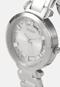 Hahlain teräksinen kello, jossa on pyöreä kellotaulu, kristallikoristeet, minimalistiset tuntimerkinnät ja kiillotettu metalliranneke.