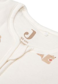 Gros plan sur l'encolure d'un vêtement pour bébé blanc avec une étiquette de taille beige indiquant "90 cm" et une partie d'une illustration d'animal brun.