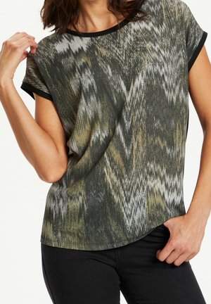 Kvinde iført T-shirt med korte ærmer med abstrakt sort, gråt og olivengrønt zigzag-mønster og sort kant ved ærmer og halsudskæring.