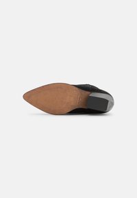 Escarpin noir en daim à talon haut avec un bout pointu et une semelle en bois. Ses caractéristiques notables incluent une texture antidérapante et une finition mate.