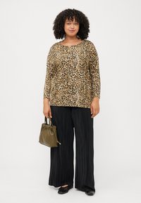 A woman with curly hair wears a leopard print top, black pleated pants, and holds an olive green handbag, standing against a white background.