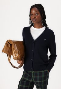 Navy knitted cardigan with a v-neckline and button closure, paired with a white t-shirt and plaid pants; carrying a glossy brown handbag.