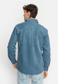 Camicia di velluto a coste blu con colletto, maniche lunghe e dettagli con bottoni. Tessuto testurizzato con righe verticali e orlo curvato.