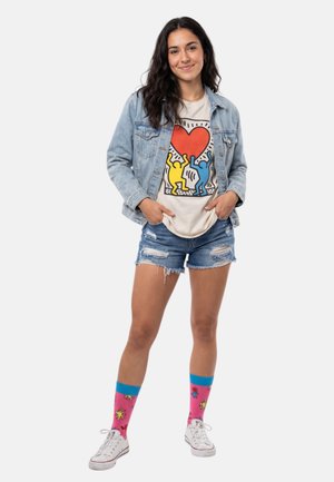 Jonge vrouw met een spijkerjack, een grafisch t-shirt met een hart en dansfiguren, gescheurde spijkershorts, kleurrijke sokken en witte sneakers.