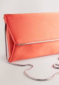 Pochette en tissu corail avec rabat triangulaire, dotée d'une bandoulière chaîne argentée et d'un design élégant. Texture lisse avec un détail de couture visible.
