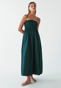 Lungo vestito verde senza maniche con un corpino arricciato e una gonna fluida. Il tessuto appare liscio e leggero. Indossate sandali piatti.
