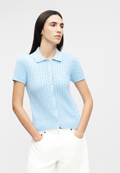 Hosszú sötét hajú nő világoskék, rövid ujjú, kötött kardigánban és fehér nadrágban, kezét zsebében tartva, jobbra néz.