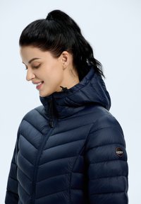 Laivastonsininen untuvatakki jossa on istuva huppu, vaakasuora tikkauskuvio, säädettävä kiristysnauha ja logopatchi vasemmassa olkapäässä.