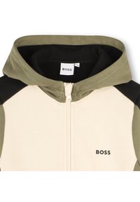 Hettegenser med glidelås i beige med grønne og svarte detaljer, med en BOSS-logo på forsiden, laget av et mykt stoff.