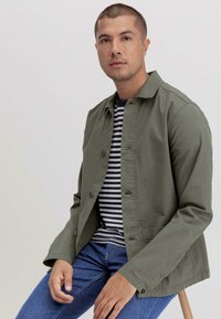 Olijfgroen lichtgewicht jack met knoopsluiting, gecombineerd met een marineblauw en wit gestreept shirt, gedragen met blauwe spijkerbroek, zittend op een kruk.