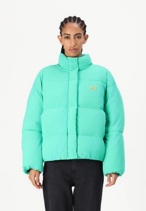 Svijetlozeleni puffer jakna s visokim ovratnikom, kopčama na gumbe i malo prevelikim krojem. Ima mali logotip na prsima.