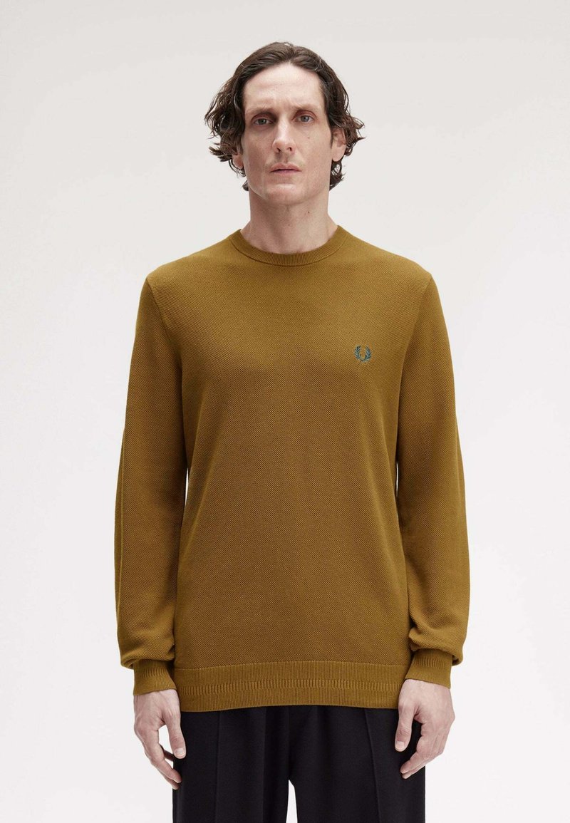 Maglione a maglia giallo senape con maniche lunghe e collo a giro. Presenta polsini e orlo a costine con un piccolo emblema sul lato sinistro del petto.