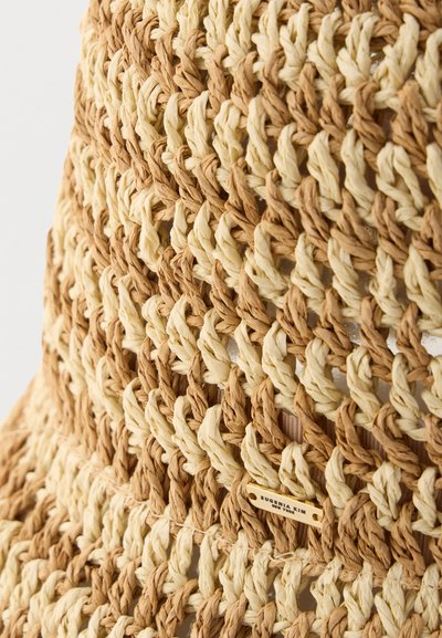 Κοντινή λήψη υφαντού μπεζ και ανοιχτό καφέ ψάθινου καπέλου με μια μικρή ορθογώνια μεταλλική ετικέτα μπροστά.