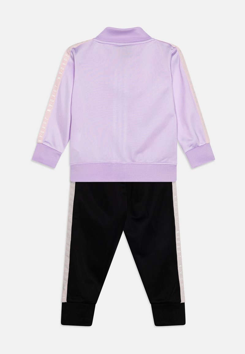 Chaqueta de chándal lila con cuello acanalado y pantalones negros con franjas laterales blancas con el texto "JORDAN". Textura de tejido suave en toda la prenda.