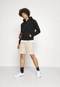 Musta huppari, beige shortsit, valkoiset sneakerit ja mustat sukat. Malli seisoo yksinkertaisen taustan edessä, kädet taskuissa, rento tyyli.
