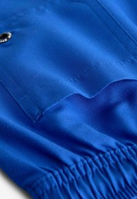 Tissu bleu vif avec une texture lisse présente un bord replié et un fronce élastique en bas. Un bouton noir est visible.