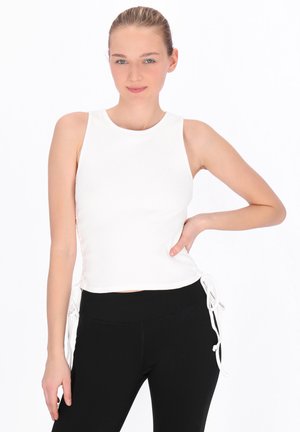 Fiatal nő szőke hajjal, fehér, oldalt megkötős ujjatlan felsőben és fekete nadrágban, egyik kezét csípőjén tartva áll.