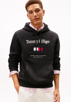 Mladý muž nosiaci čiernu mikinu Tommy Hilfiger cez pruhované tričko s bielymi nohavicami, stojaci s jednou rukou v vrecku.