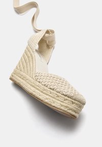 Beige espadrillekilklädd sandalett med vävd ovandel, rund tå och flätad jutecula. Har en mjuk tygrem runt ankeln.
