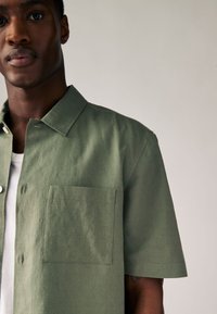 Kortærmet, olivengrøn skjorte med knapper i let stof, der har en brystlomme og en klassisk krave, og viser en jævn tekstur.