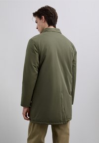 Olivengrøn frakke med lange ærmer og krave, glat tekstur og sidelommer, med en lige pasform og slids bagpå.