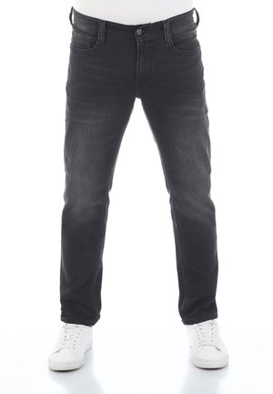 Mann trägt eng anliegende schwarze Jeans und weiße Sneakers, steht vor weißem Hintergrund.