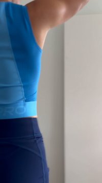 Person iført blå atletisk top og mørke leggings, løfter armene indendørs mod en ensfarvet lys væg.