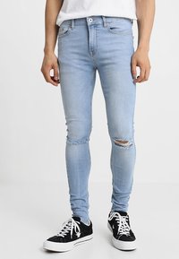 Mies, joka on pukeutunut vaaleansinisiin revittyihin skinny-farkkuihin ja mustiin matalavartisiin tennareihin, joissa on valkoiset nauhat, seisoo valkoista taustaa vasten.