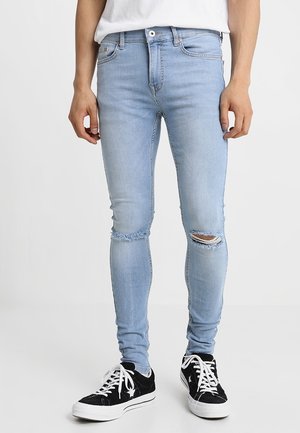 Man som bär ljusblå slitna slim-fit jeans och svarta låga sneakers med vita snörningar, stående framför en vit bakgrund.