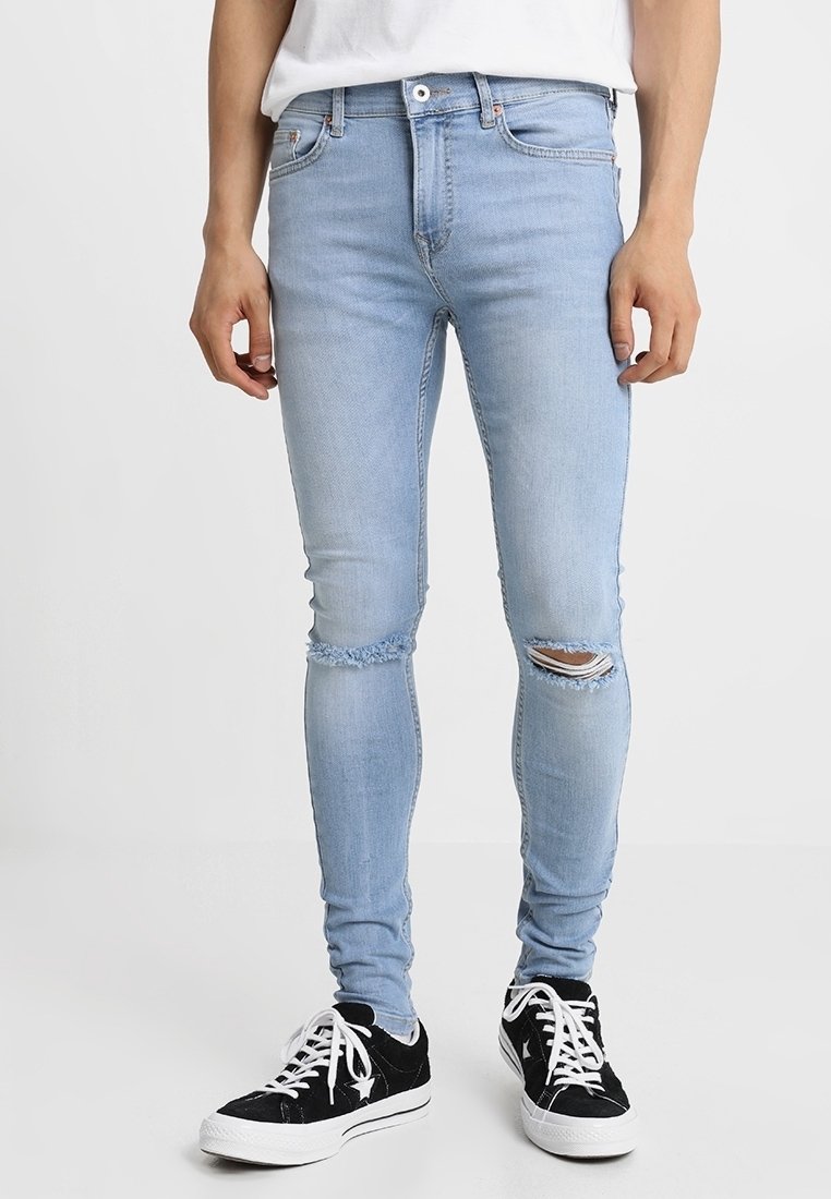 Mies, joka on pukeutunut vaaleansinisiin revittyihin skinny-farkkuihin ja mustiin matalavartisiin tennareihin, joissa on valkoiset nauhat, seisoo valkoista taustaa vasten.