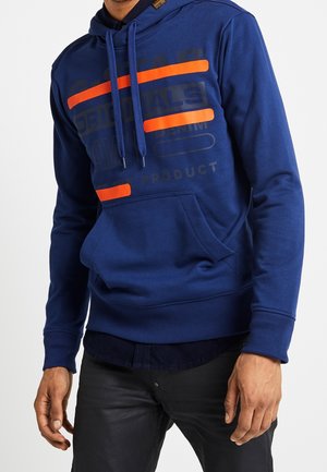 Mand iført en blå hættetrøje med orange striber og sorte bukser, stående med hænderne afslappet ned langs siden mod en hvid baggrund.