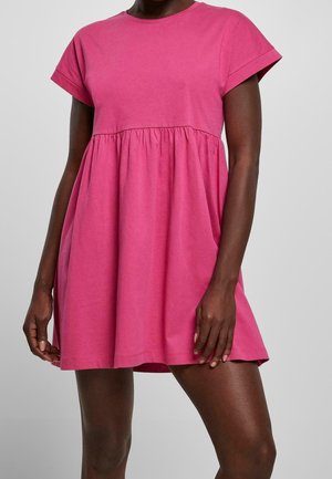 Femme portant une robe rose à manches courtes, ample, avec une taille froncée et un ourlet au-dessus du genou, sur un fond gris uni.