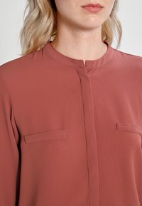 Blouse couleur rouille avec un col rond, une fermeture à boutons à l'avant et deux poches poitrine plaquées. Fabriquée en tissu léger et lisse avec un léger éclat.