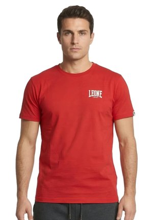 Un homme portant un T-shirt rouge Leone 1947 et un pantalon foncé se tient devant un fond blanc.