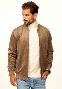 Braune Wildleder-Bomberjacke mit Rippbündchen und -saum, Reißverschluss, die über einem cremefarbenen Rollkragenpullover getragen wird. Hellblaue Jeans runden das Outfit ab.