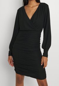 Robe noire en tissu texturé, avec des manches longues, un décolleté en V profond et des détails froncés sur les côtés pour un effet ajusté.