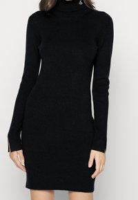 Robe noire côtelée avec un col roulé haut et des manches longues. Présente des fentes latérales à l'ourlet et un petit détail de logo au niveau du col.