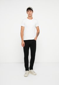 Fehér pamut póló, slim fit fekete farmernadrág, bézs tornacipő és szürkesapka. A modell egy sima fehér háttér előtt áll.
