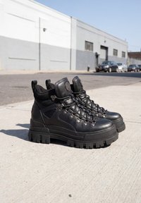 Paire de bottes de randonnée en cuir noir avec semelles épaisses et lacets, posées sur un trottoir urbain ensoleillé avec des voitures en arrière-plan.