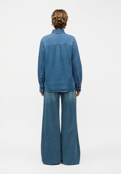 Άτομο με σγουρά μαλλιά που φοράει μπλε τζιν πουκάμισο και τζιν παντελόνι με φαρδιά μπατζάκια, γυρισμένο προς τα πίσω μπροστά σε ένα απλό φωτεινό φόντο.