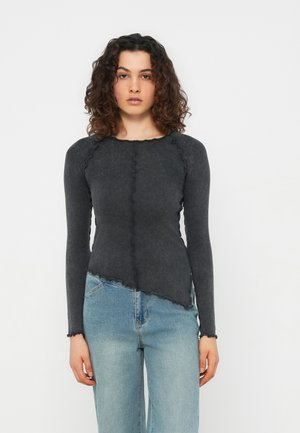 Jeune femme aux cheveux bouclés portant un haut ajusté à manches longues gris foncé et un jean taille haute bleu clair, debout devant un fond blanc.