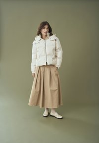 Balta pūkinė striukė su kapišonu ir spaudžiamomis kišenėmis, derinama su bežine klostuota sijonu. Modelis vilki baltus loafer stiliaus batelius ir raštuotas pėdkelnes.