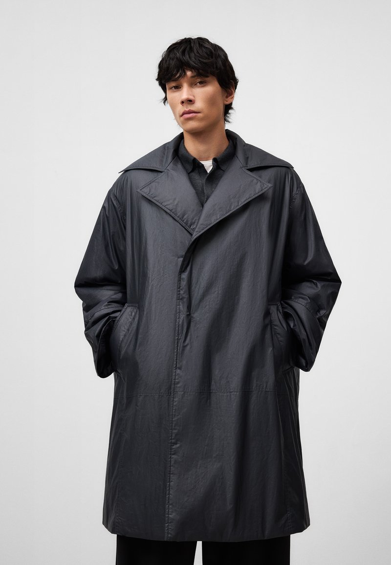 Długi, ciemnoszary płaszcz trench wykończony z lekkiego, gładkiego materiału. Posiada duży kołnierz, boczne kieszenie oraz prosty krój. Minimalistyczny design.
