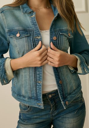 Femme boutonnant les boutons avant d'une veste en denim bleu délavé portée sur un débardeur blanc côtelé avec un jean en denim assorti.
