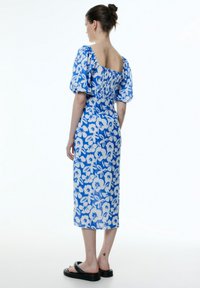 Maxikleid in blauer Blumenoptik mit Puffärmel, gerüschtem Rücken und taillierter Silhouette. Flache schwarze Sandalen ergänzen das Outfit.