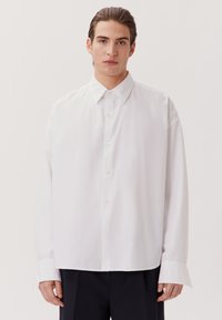 Camicia bianca a maniche lunghe con colletto a punta, caratterizzata da una vestibilità ampia, chiusura frontale con bottoni e polsini larghi, realizzata in tessuto morbido e leggero.