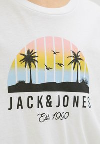 Valkoinen puuvillapaita, jossa on värikäs grafiikka palmuista ja linnuista auringonlaskun väriarvoista taustaa vasten. "JACK & JONES" painettuna alle.