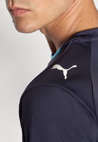 Camisa desportiva azul-marinho com tecido texturizado, apresentando um colarinho turquesa e um logótipo da Puma branco no ombro. Close-up mostrando o detalhe do ombro.