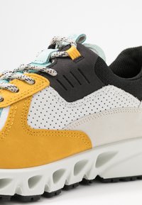 Sneakers med en blandning av material: gul mocka, svart nät och vit perforerad läder. Har texturerade snörningar och en formad vit sula.
