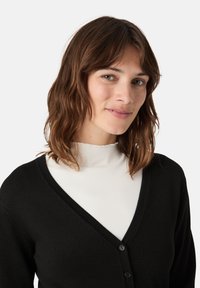 Černý kardigan s výstřihem do V, vyrobený z měkkého pleteného materiálu. Obsahuje dva knoflíky a je vrstvený přes bílý žebrovaný rolák.